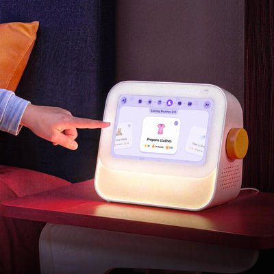 A young girl sits on a bed next to a bedside table, pointing at the Cozyla Routie, the smart kids' routine machine, displaying the Evening Routine interface with soothing night light
