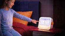 A young girl sits on a bed next to a bedside table, pointing at the Cozyla Routie, the smart kids' routine machine, displaying the Evening Routine interface with soothing night light